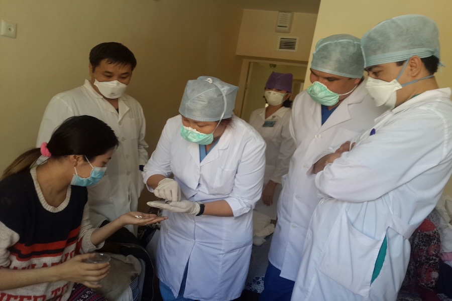 A patient stands at a counter while a medical professional in a white coat writes in an office.