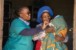 A healthcare worker gently checks on a bundled newborn while the mother smiles and holds the baby close, wrapped in a green blanket.