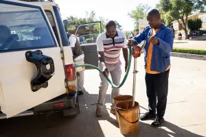 Two men refueling a white truck from a bucket with a hose and pump.