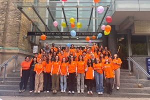 A large group of students posing on a staircase, wearing orange shirts and tossing orange balloons.