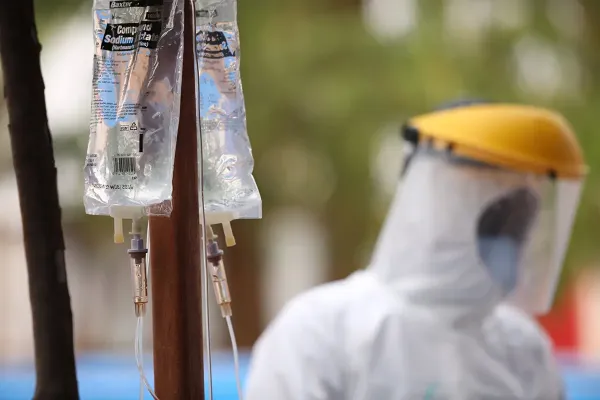 In the foreground is a clear medical bag hooked up to drip. In the background, there is a blurred health care worker wearing white protective equipment and a mask.