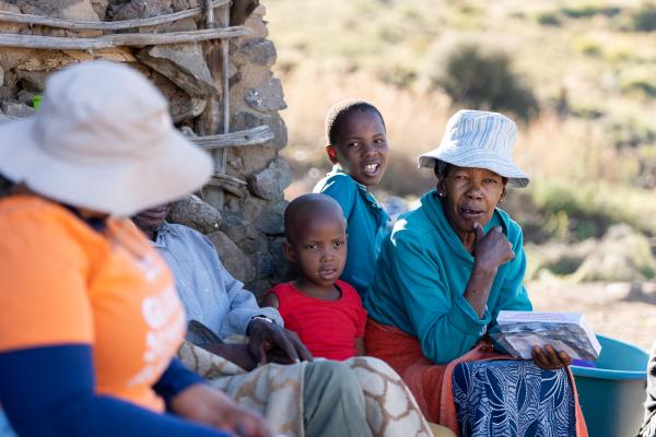 A group of four people, including an elderly woman, two children, and a health worker wearing an orange PIH shirt, sit together outdoors. The elderly woman, wearing a hat, is engaged in conversation, holding a book in her hands, while the health worker listens.
