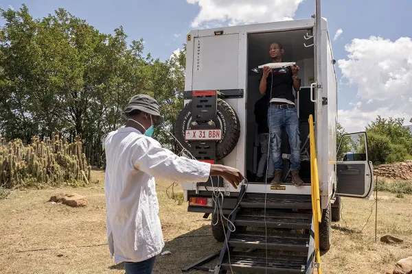 A man stands in the back of a white utility truck while another man on the ground is handling a cable. They are in a dry, grassy area with trees and cacti visible under a partly cloudy sky.
