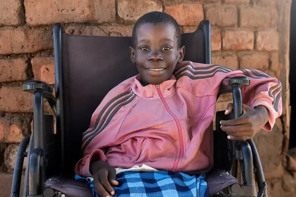 A young girl wearing a pink shirt sitting in a wheelchair looks at the camera smiling, with a red brick wall behind her.