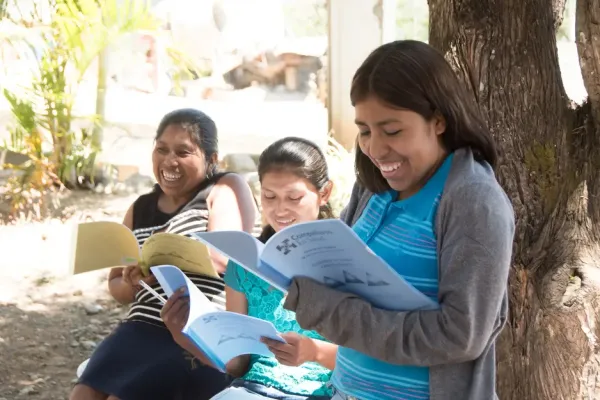 Three smiling women reading booklets while seated under a tree.