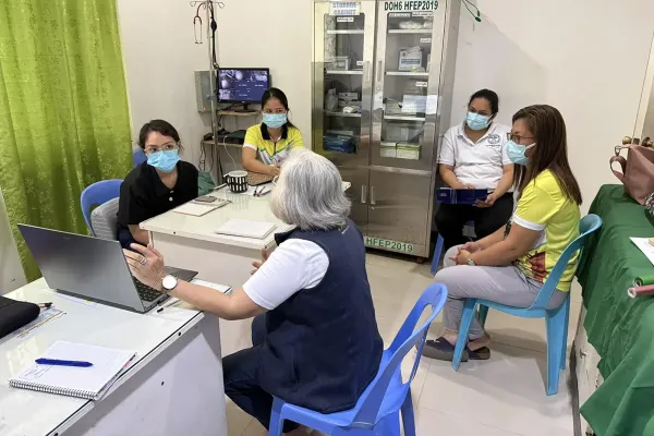 Four women wearing medical masks sit in blue plastic chairs around a white table are given instruction by a person gesturing at a computer.