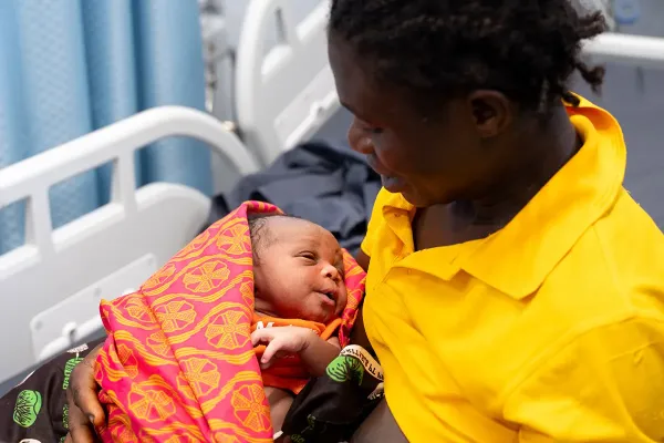 A mom wearing a yellow shirt holds her new born baby swaddled in a red and orange patterned blanket.
