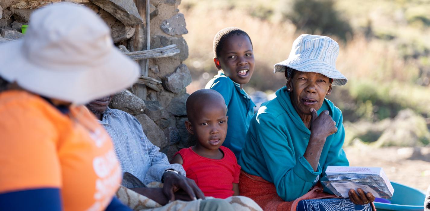 A group of four people, including an elderly woman, two children, and a health worker wearing an orange PIH shirt, sit together outdoors. The elderly woman, wearing a hat, is engaged in conversation, holding a book in her hands, while the health worker listens.