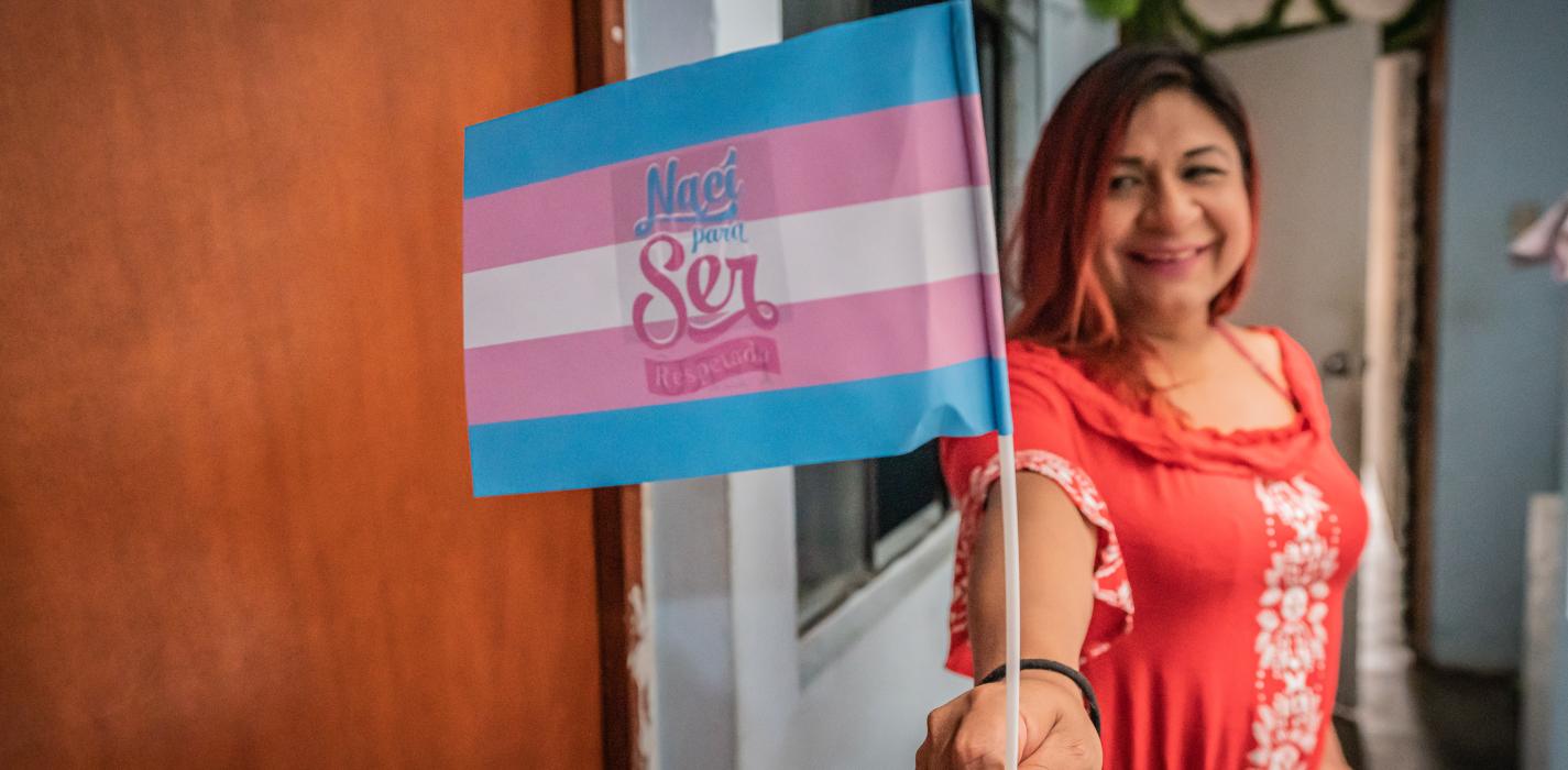 Brenda Mijahuanca smiles while holding up a tricolour white, blue, and pink flag, expressing joy.