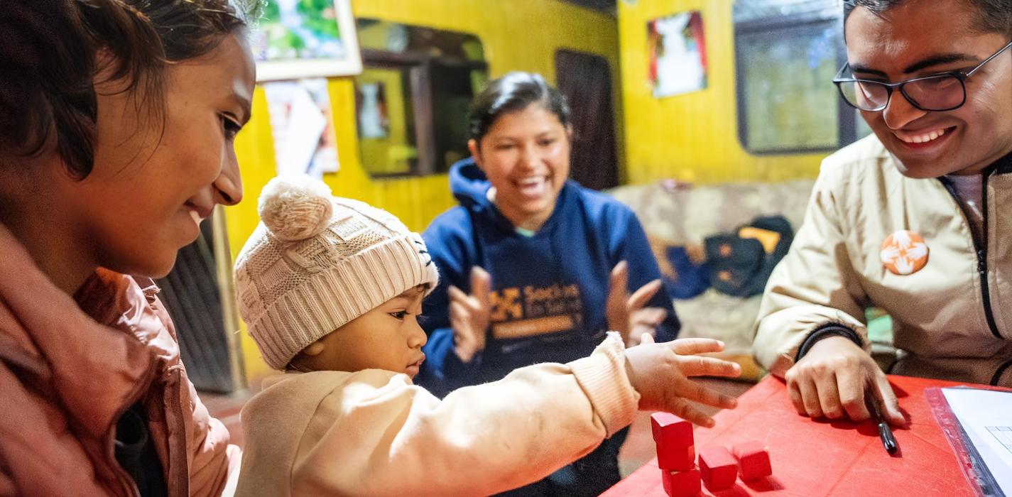 A baby in a white hat reaches for red blocks on a table, surrounded by three smiling adults in a brightly lit room.