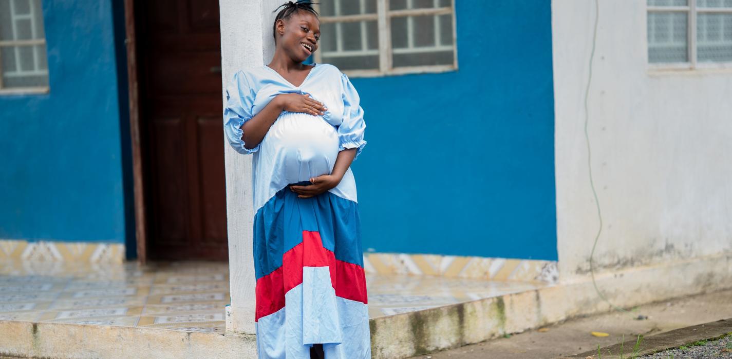 A pregnant woman stands outside a blue and white building, smiling warmly as she rests her hands on her belly. She wears a long blue dress with red and white accents and sandals, leaning slightly against a white column. The scene feels calm and hopeful.