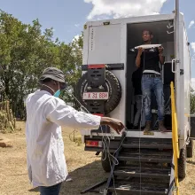 A man stands in the back of a white utility truck while another man on the ground is handling a cable. They are in a dry, grassy area with trees and cacti visible under a partly cloudy sky.