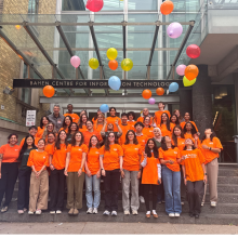 A large group of students posing on a staircase, wearing orange shirts and tossing orange balloons.