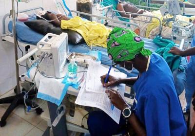 A doctor wearing scrubs is leaned over equipment, writing a patient report on a clipboard. A hospital setting visible in the background.