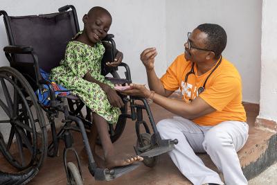 A doctor wearing an orange PIH t-shirt sits on a step talking to Regina, who is sitting in her wheelchair.