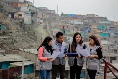 Four people in grey vests stand together reviewing documents in a densely populated hillside neighbourhood.