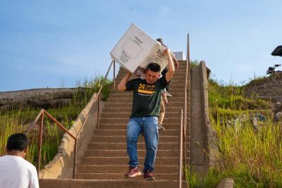 A man is carefully descending a set of concrete stairs, carrying a heavy box over his shoulders, with a focused expression as he navigates the steps.