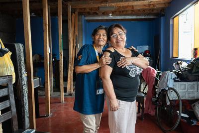 Two women stand in a modest home, one behind the other with hands on her shoulders in a supportive gesture, a wheelchair visible nearby.