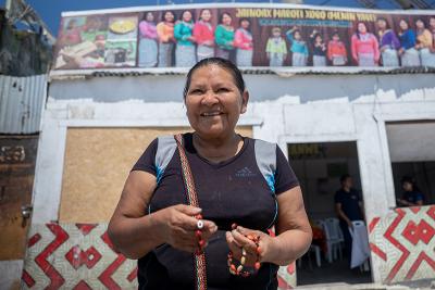 A smiling woman holds a beaded bracelet outside a community building.