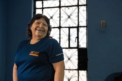 A smiling woman wearing a Socios En Salud T-shirt stands inside a blue-walled room beside a decorative metal door.