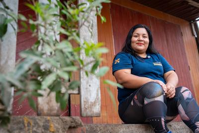 A woman wearing a Socios En Salud shirt sits on the steps outside a wooden building, with leafy plants in the foreground