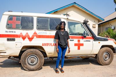 A woman wearing a black shirt and blue jeans stands in front of an ambulance on a sunny day.