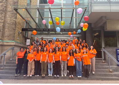 A large group of students posing on a staircase, wearing orange shirts and tossing orange balloons.