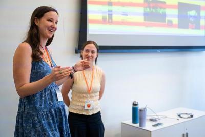 A woman in a blue dress speaking and gesturing with her hands, standing beside another woman in a white shirt.