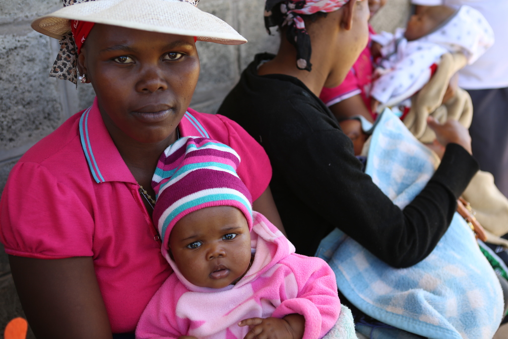 A mother holds her young daughter sitting outside a clinic with other women and babies in the background