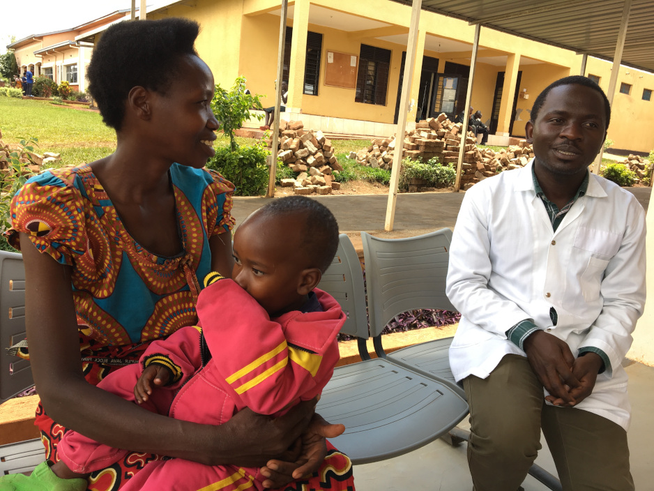 A mother holds her two year old child on her lap while sitting and speaking with a nurse on a bunch outside the clinic