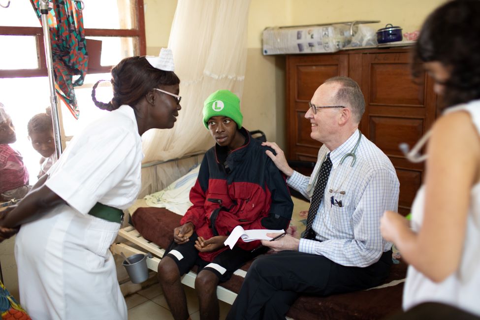 Dr. Paul farmer sits on a bed with a young male patient as they talk with a nurse